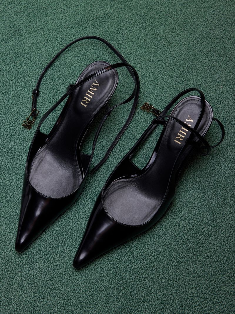 Pair of black AMIRI pointed-toe slingback high heels on a textured green surface, showcasing modern fashion footwear.