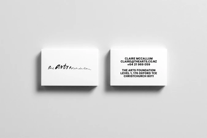 Two white business cards resting on a pale grey surface, showing a handwritten brand logo on one and contact details on the other.