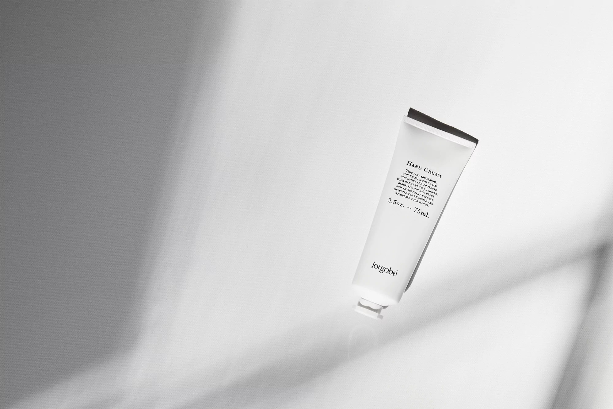 A tube of hand cream lies on a white surface with light and shadow creating a minimalist, high-key beauty product shot.