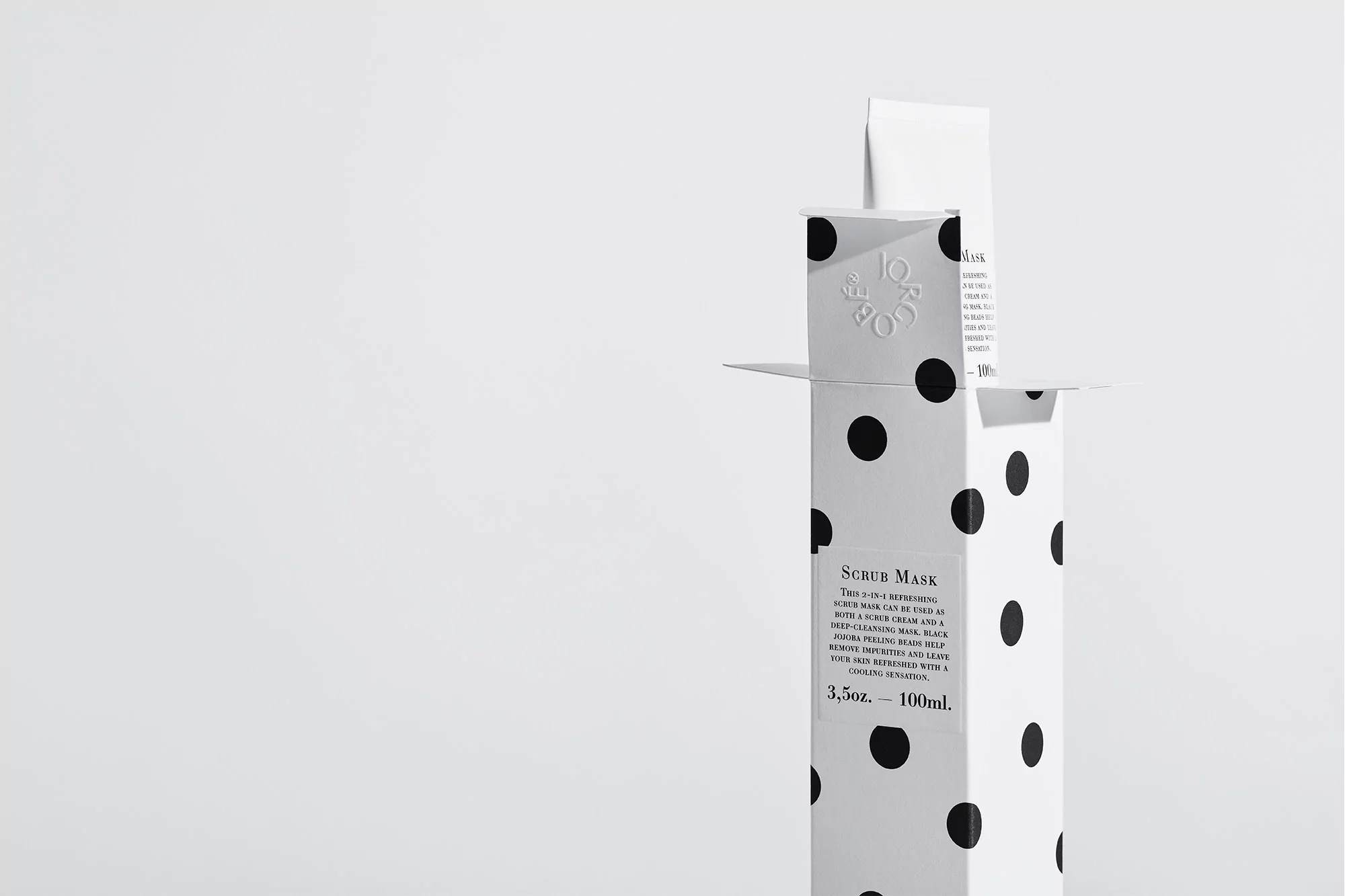 Close-up of a scrub mask in a white cardboard box with black polka dots against a stark white backdrop, product on display.