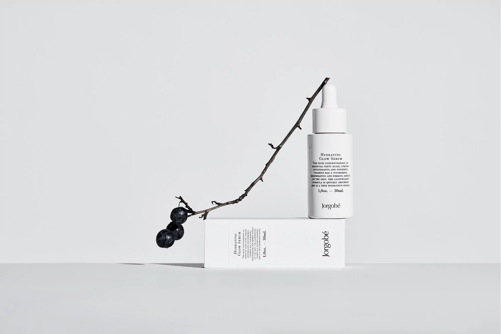 Cosmetic serum bottle with dropper and brand label next to a branch with dark berries, set on a neutral surface and backdrop.