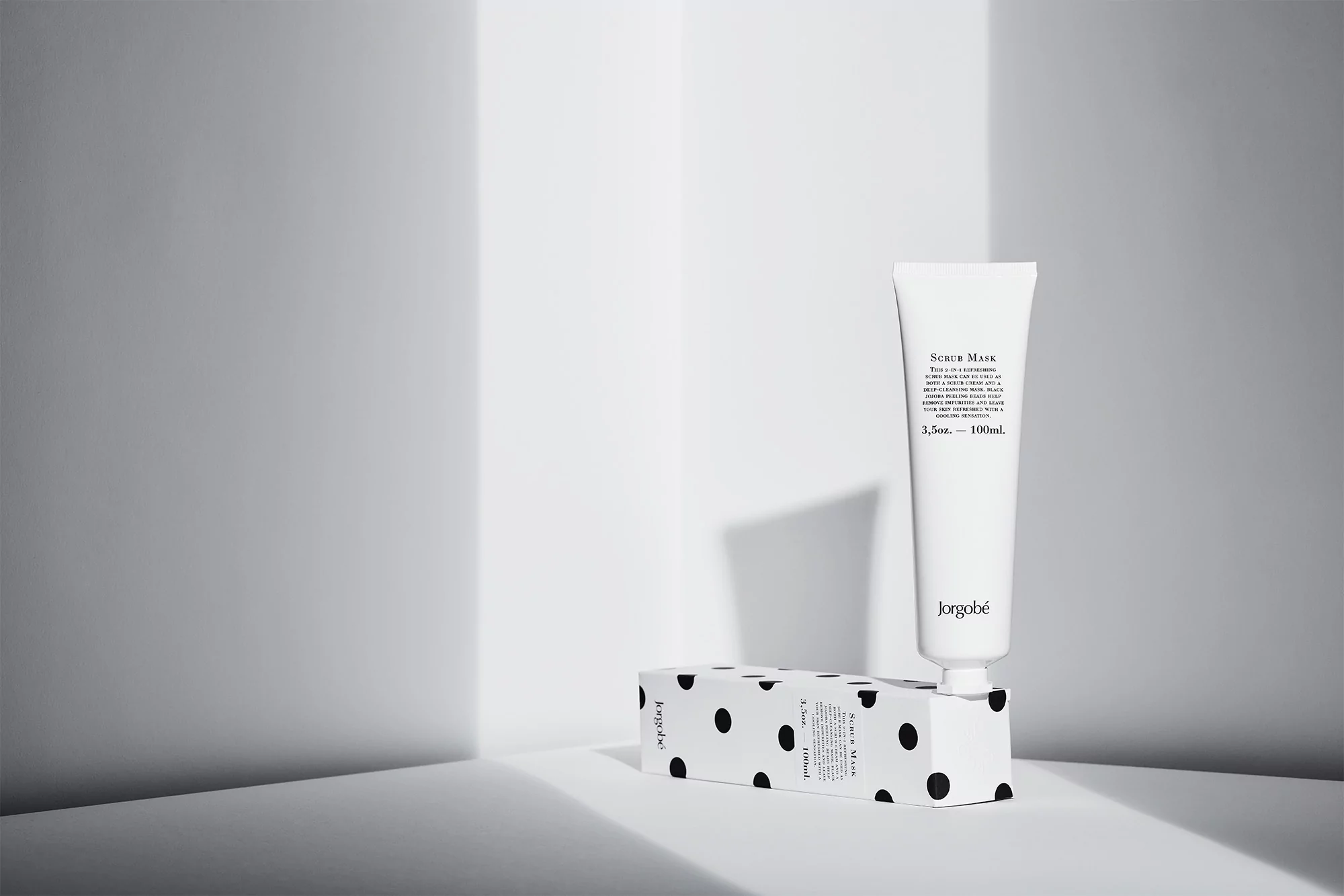 A white tube of Jorgobé scrub mask is displayed against a patterned box and a minimalist gray background in a beauty still life.