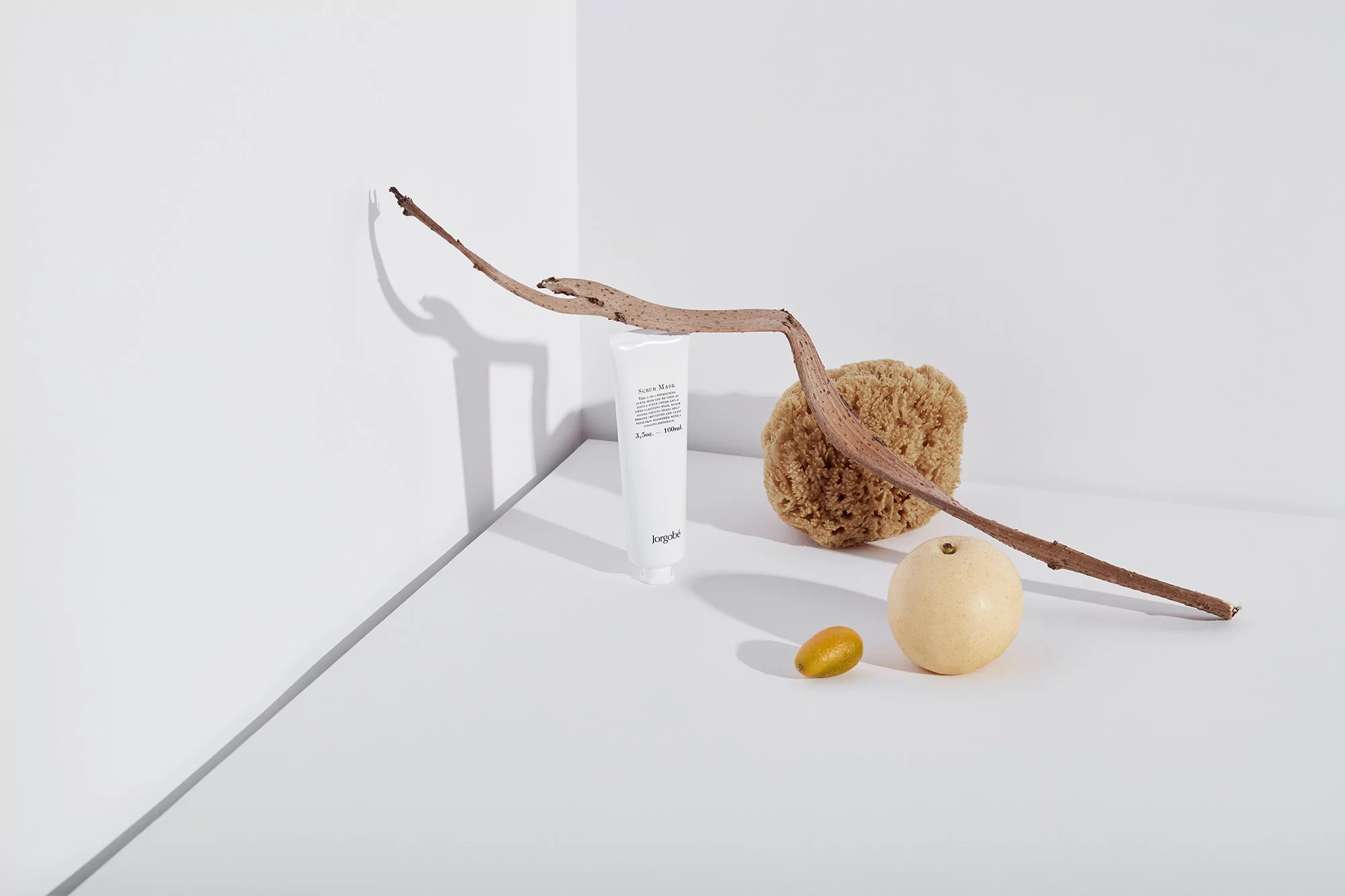 Minimalist still life featuring Jorgobe cream, sea sponge, branch, pear, and kumquat against a white corner backdrop.