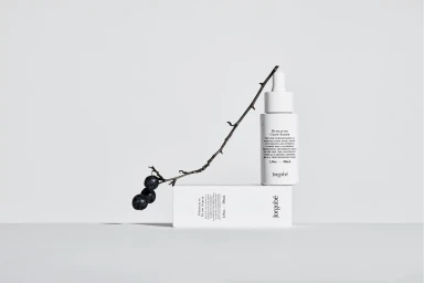 White skincare serum bottle resting on a white box with a thin twig bearing dark berries draped across them against a neutral background.