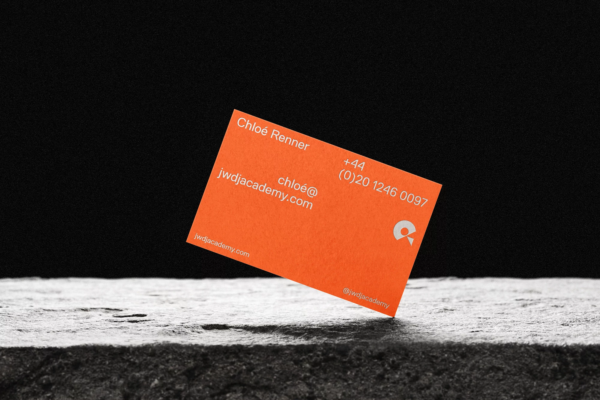 Close-up of an orange business card for jwdjacademy.com resting on a textured gray stone surface, set against a dark background.