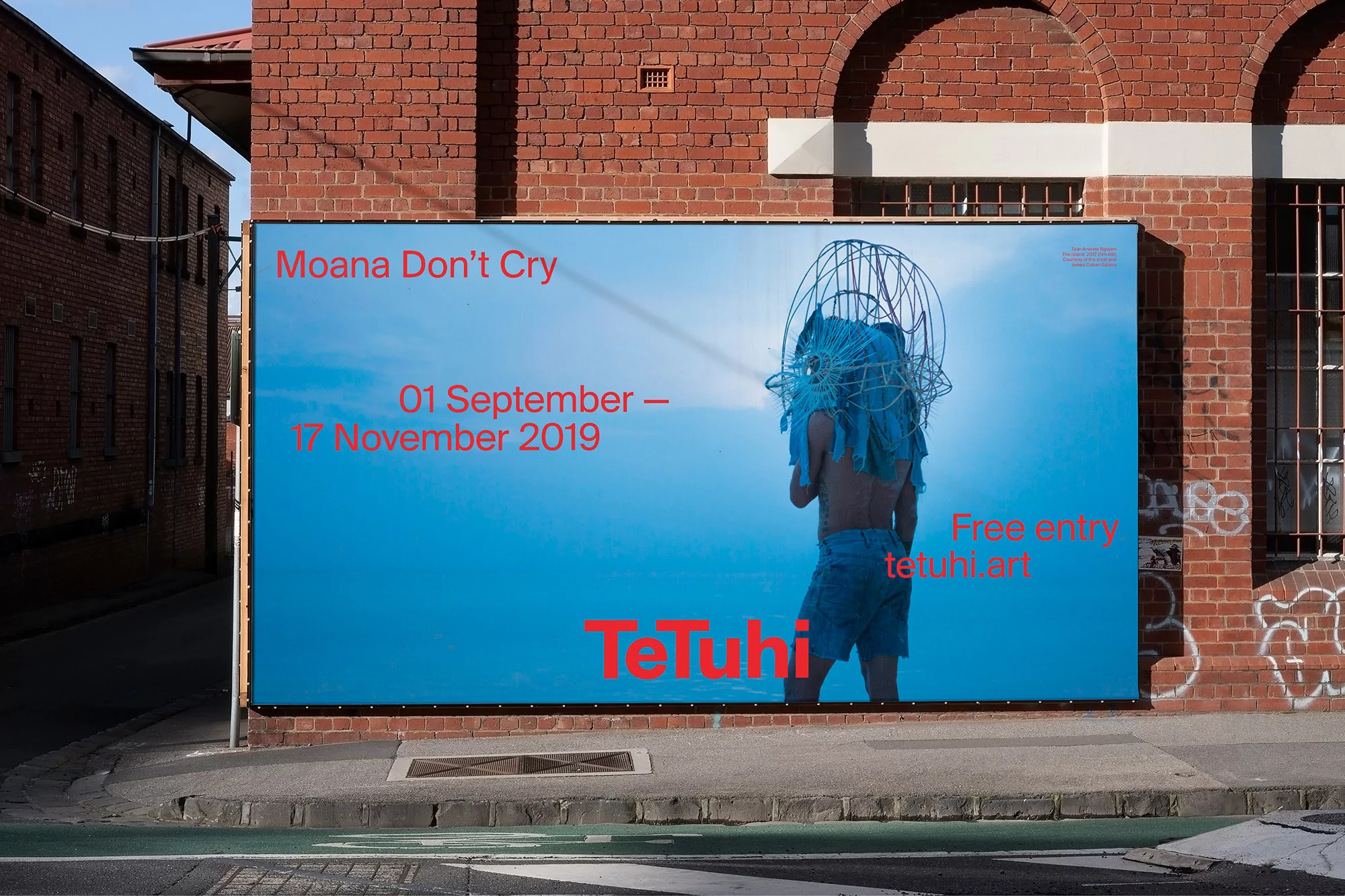 Outdoor view of a brick building displaying a large poster of a woman with sculptural headpiece, promoting an art exhibition.