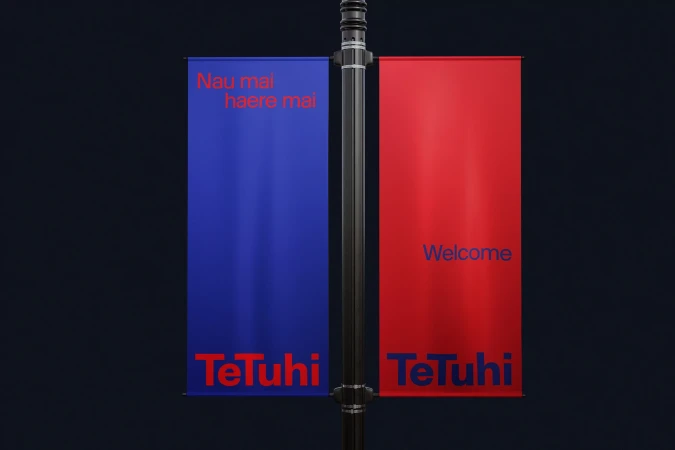 Two vertical flags on a black pole displaying brand logo against a dark background, one blue with Maori text and one red with Welcome.