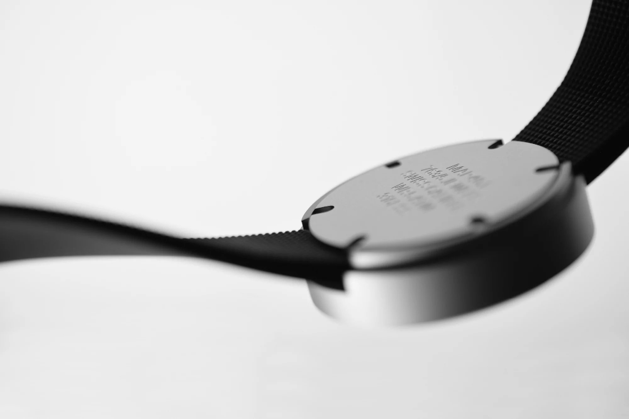 Close-up of a black watch band connected to a round, silver watch face with engraved text, set against a bright white background.