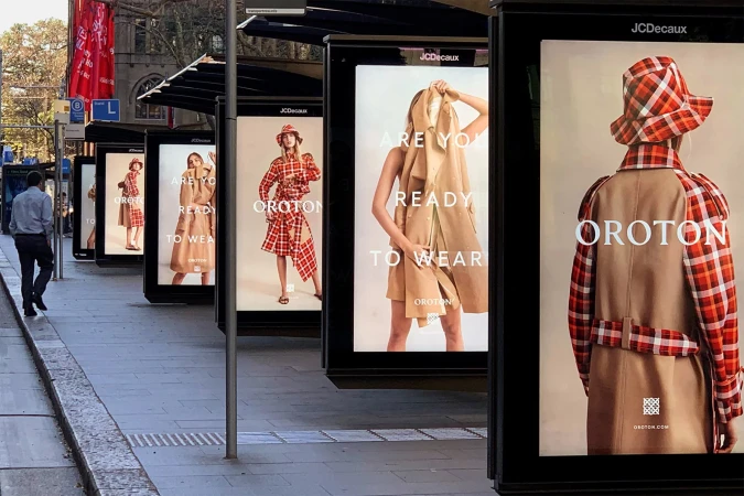 Multiple fashion ads at a bus stop feature a fair-skinned woman wearing an Oroton red plaid hat and coat, along with a tan dress.