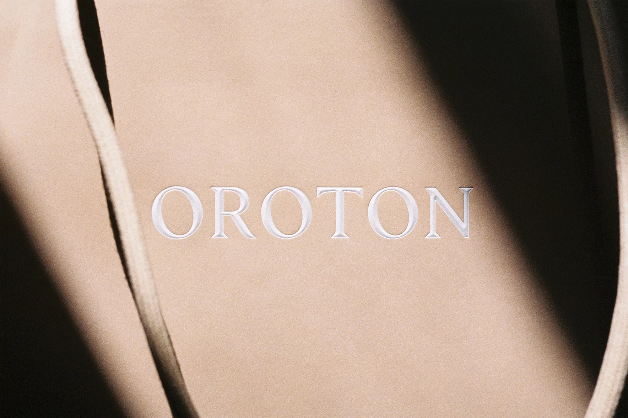 Close-up of a tan-colored fabric bag with drawstring and the brand name OROTON embossed in white, shadows and light visible.
