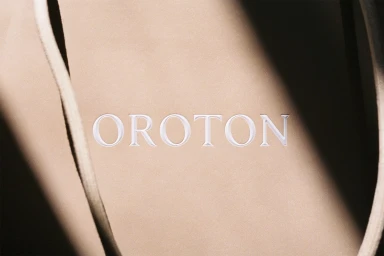 Close-up of debossed Oroton brand logo on smooth beige leather surface with subtle diagonal light and shadow.