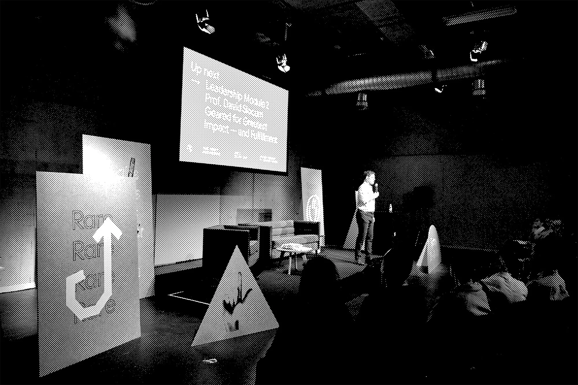 Presentation in a dark lecture hall featuring a speaker on stage in front of a screen, and audience members sitting in the foreground.