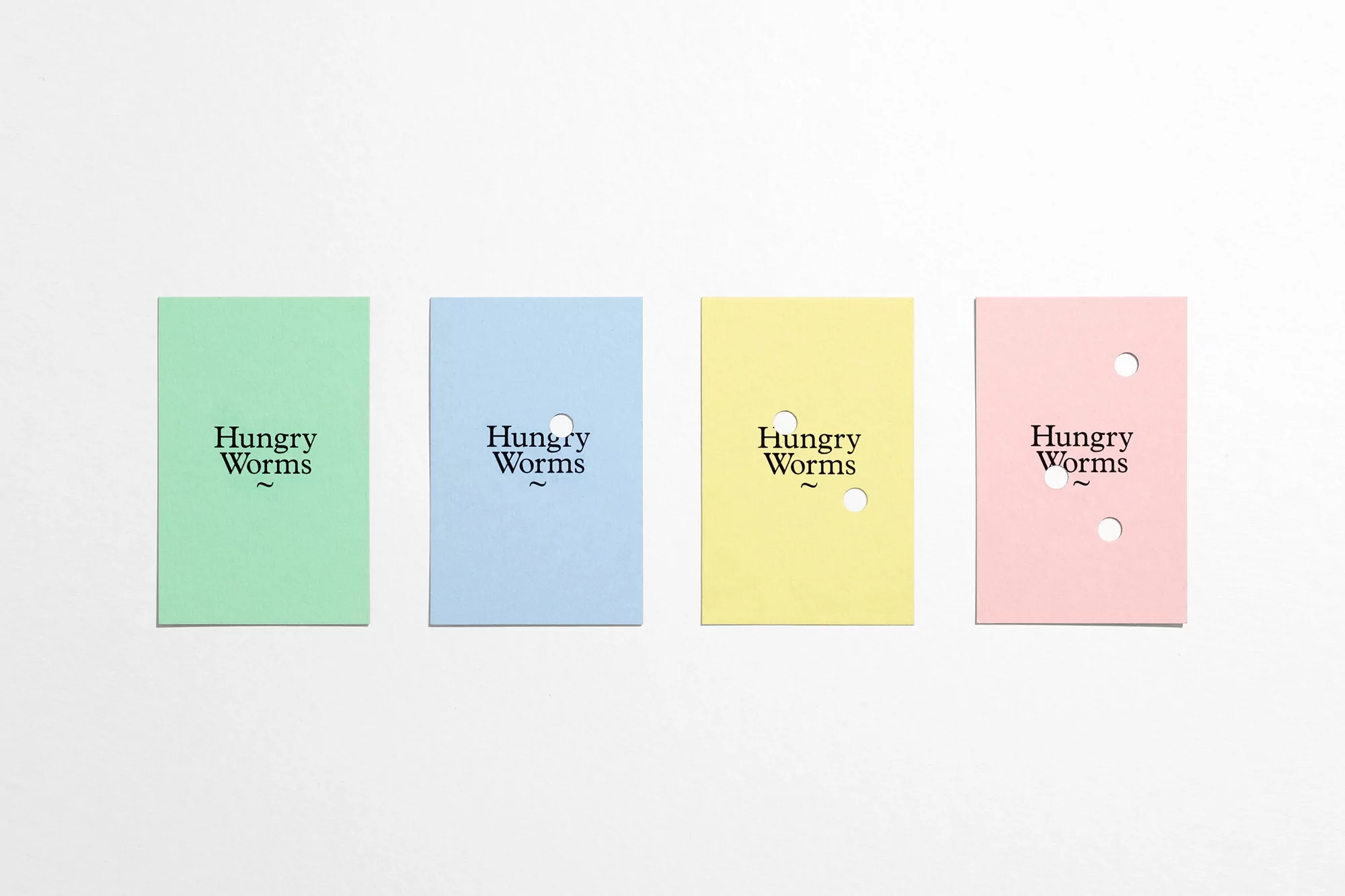 Four pastel-colored cards with 'Hungry Worms' text and die-cut holes arranged on a white surface, creating a clean and minimalist composition.