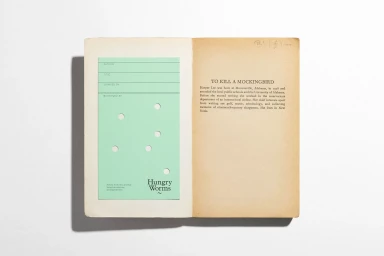 Open book displaying a green library book card with cutout holes and Hungry Worms branding against a plain white background.