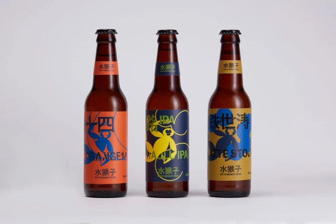 Three brown glass beer bottles with colorful labels sit against a white backdrop, each featuring unique stylized designs and text.