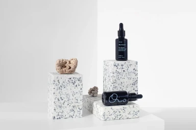 Black glass dropper bottles displayed on geometric terrazzo blocks alongside small rough stones against a clean white background