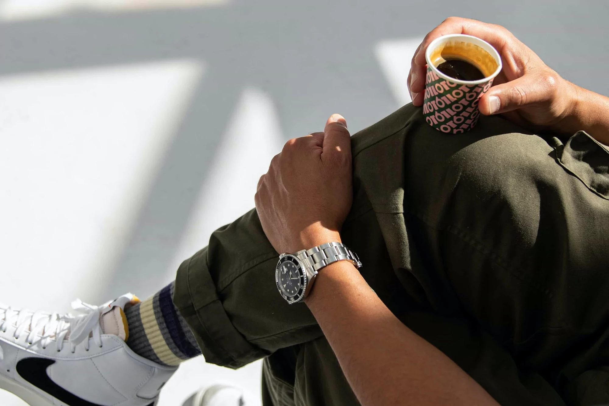 Person sits with coffee cup, showcasing wristwatch, sneakers, and olive-green attire in a sunlit indoor setting.