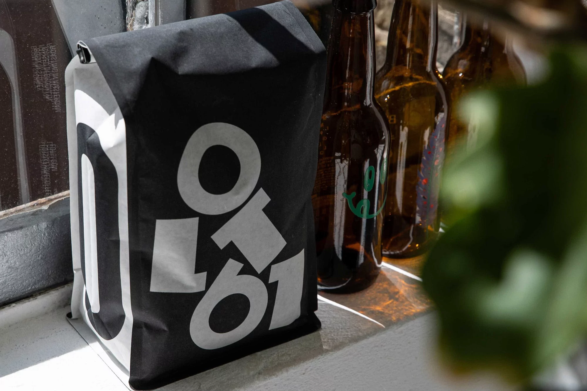 Black paper shopping bag with white logo text sits on a white windowsill with empty amber beer bottles in the background.