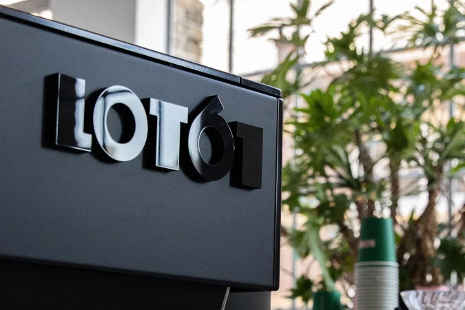 Close-up of the 'LOTO61' logo on a dark surface, with blurred plants and window light in the background, suggesting an indoor cafe setting.