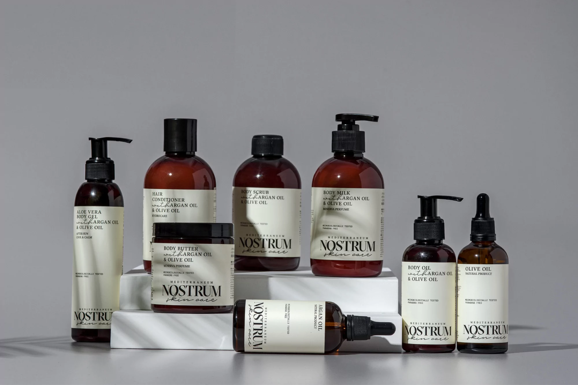 Bottles and jars of Mediterranean skincare products by Nostrum, including body oil, body butter, scrub, and milk, displayed on white blocks.