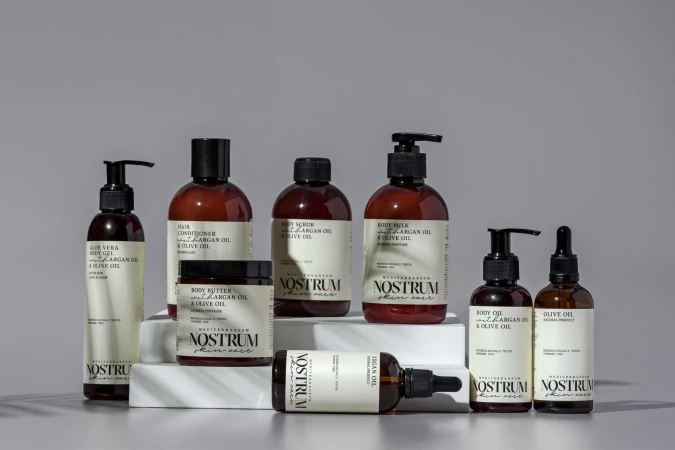 Bottles and jars of Mediterranean skincare products by Nostrum, including body oil, body butter, scrub, and milk, displayed on white blocks.