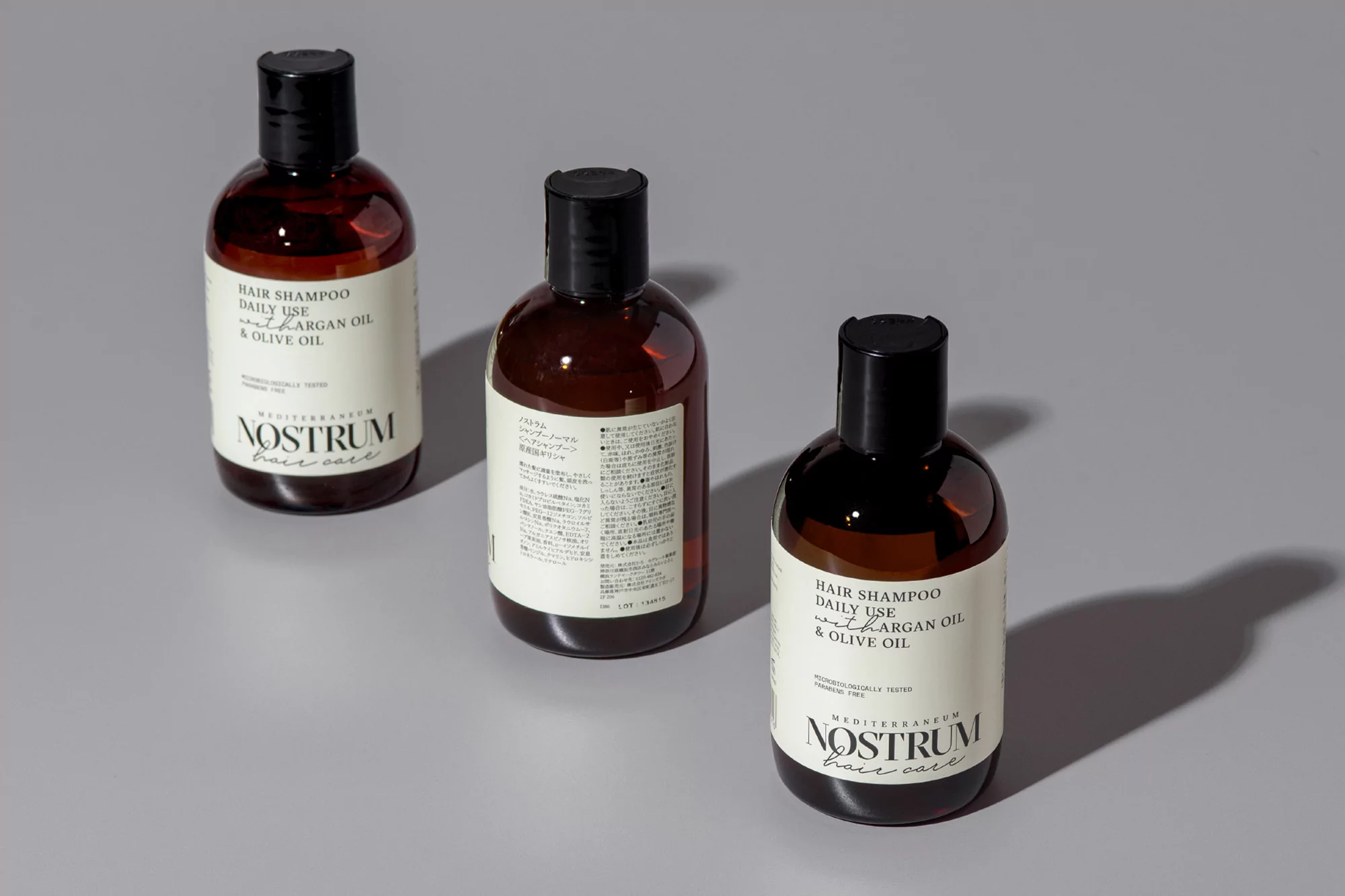 Three brown, translucent plastic bottles of hair shampoo are positioned on a light grey surface in a studio product shot.