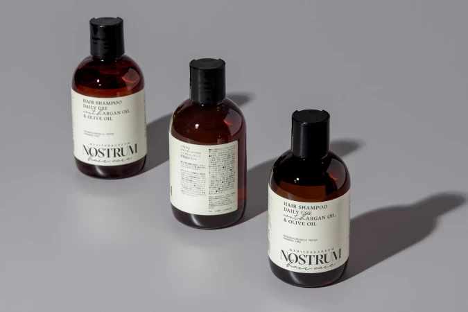 Three brown, translucent plastic bottles of hair shampoo are positioned on a light grey surface in a studio product shot.