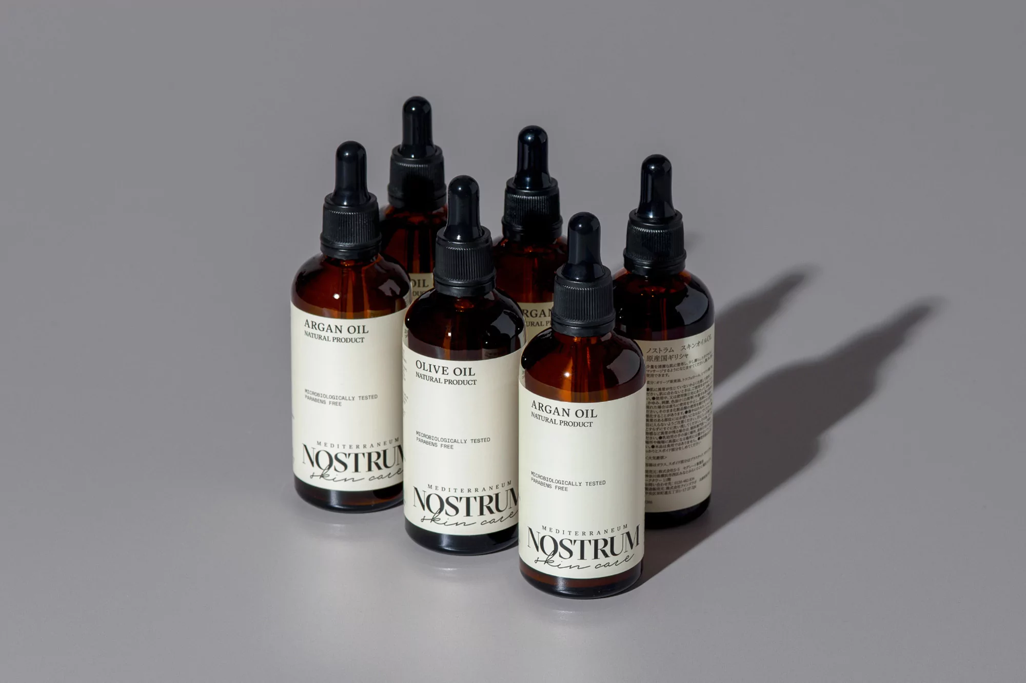 Several amber glass bottles with black dropper caps, labelled 'Argan Oil' and 'Olive Oil,' stand on a light gray surface, casting shadows.