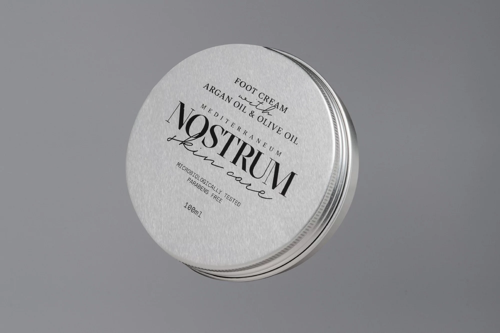 Silver tin of Nostrum skin care foot cream with argan oil and olive oil, isolated on a grey studio background, angled view.