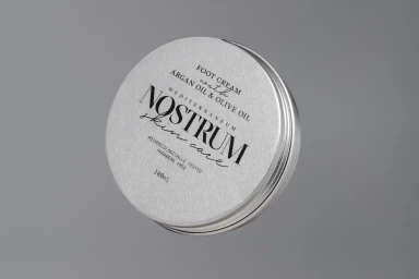 Close-up of a silver skincare tin jar for Nostrum foot cream angled against a neutral grey studio background.