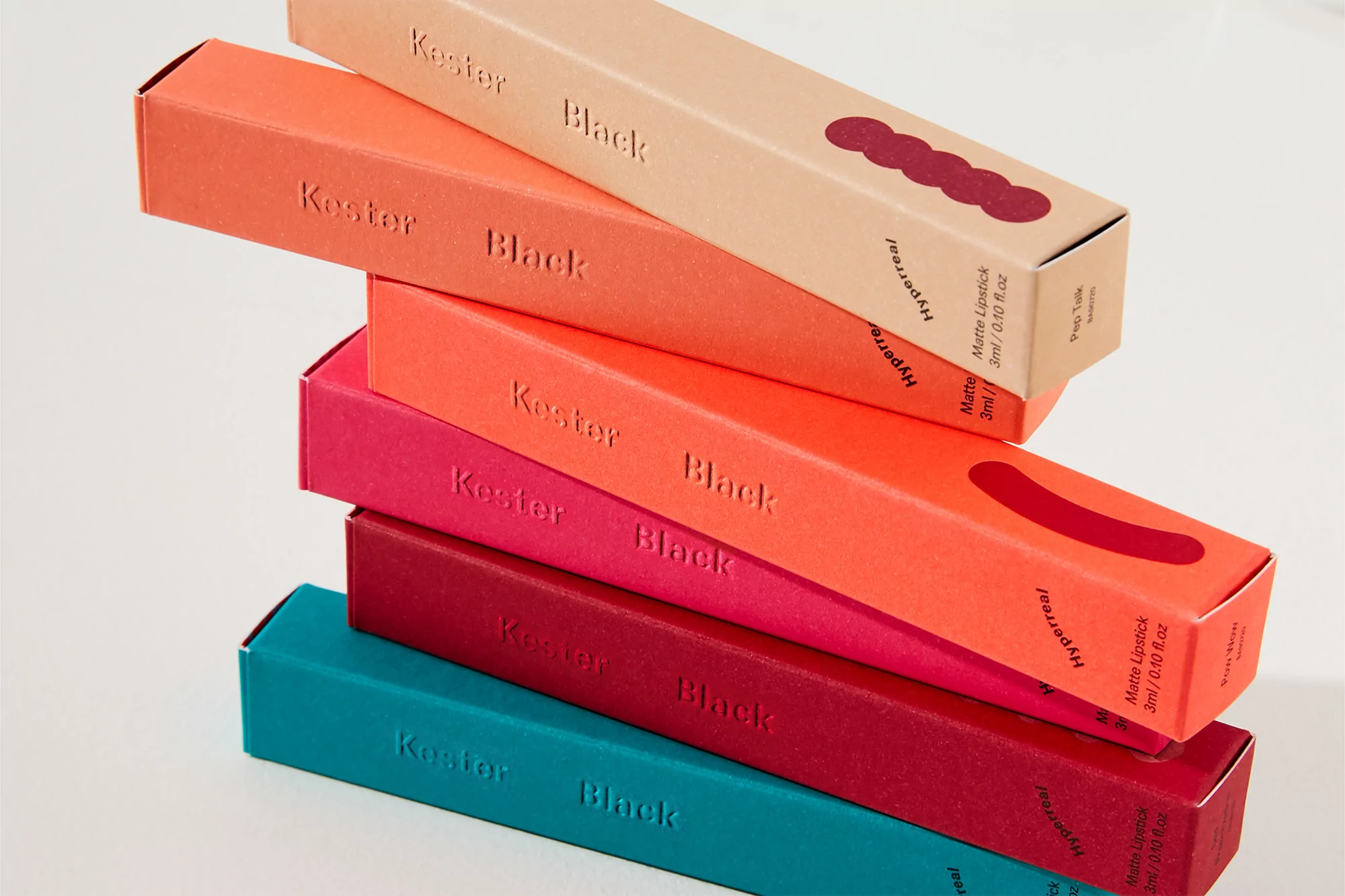 Stack of colorful Kester Black Hyperreal matte lipstick boxes in shades of teal, red, coral, and orange against a white background.
