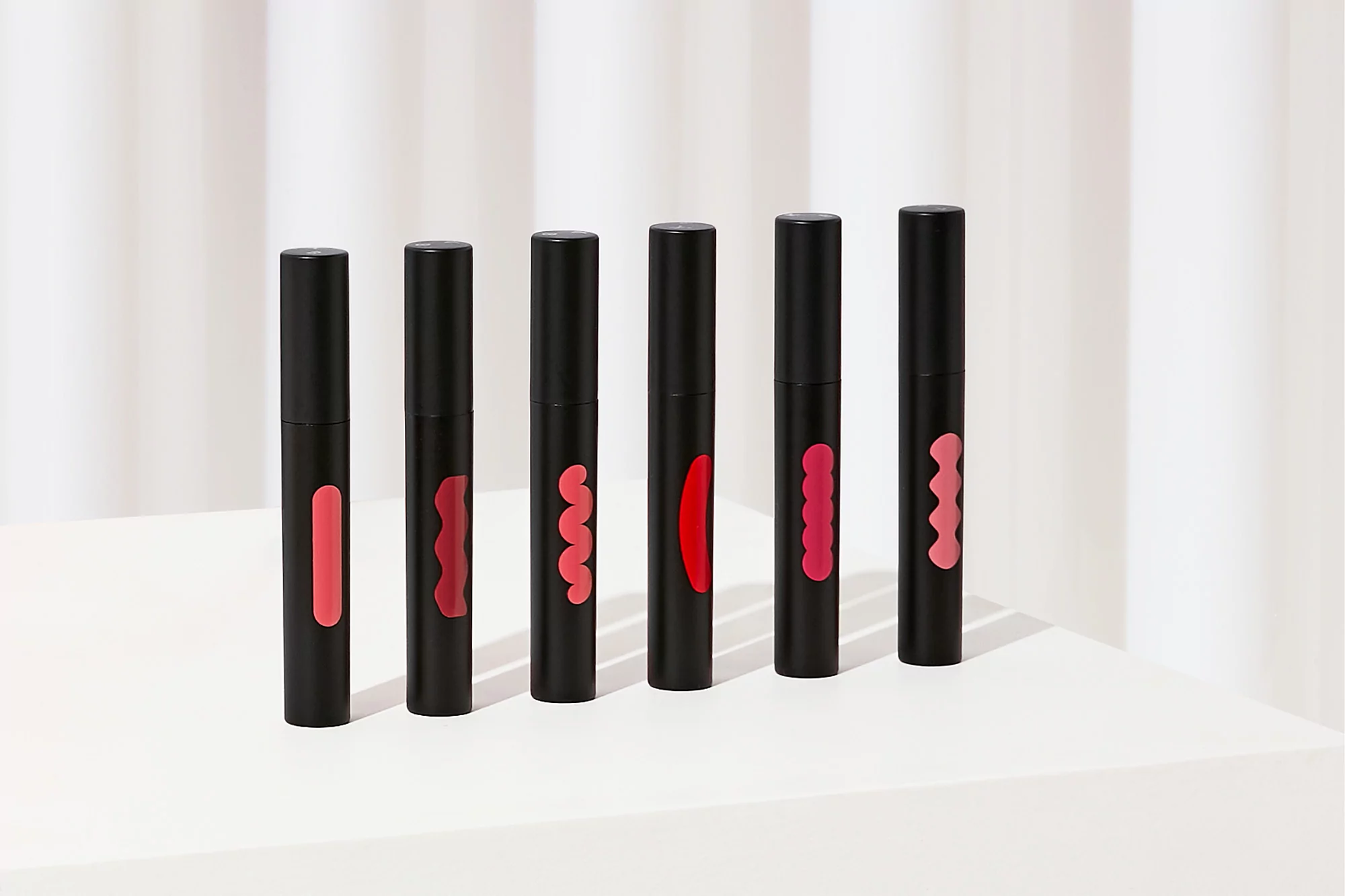 Cosmetics still life featuring six black tubes of lip gloss on a white surface against a bright white curtain background. Beauty product shot.