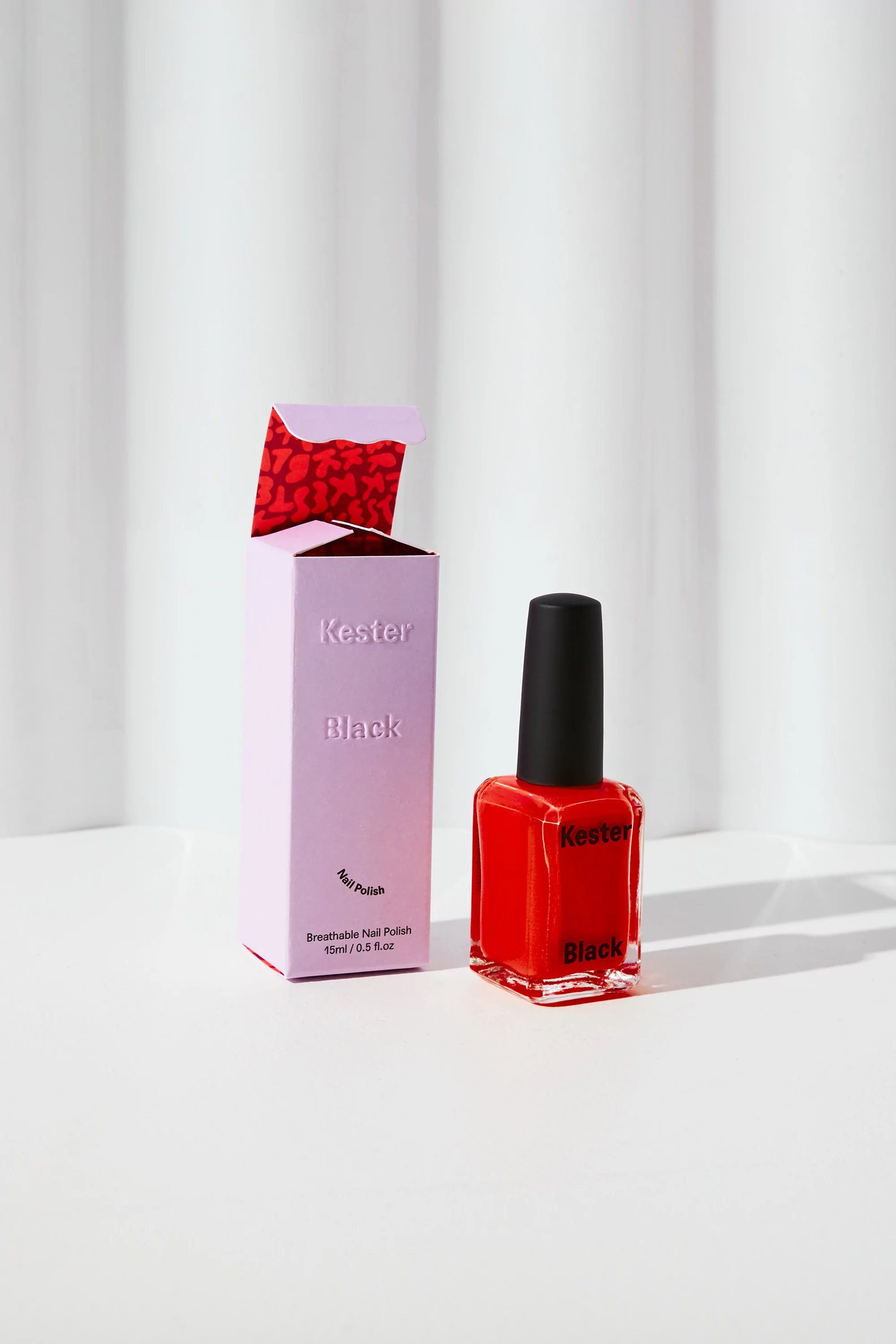 Studio shot of a red nail polish bottle with a black cap next to a lilac box on a white surface with a blurred white backdrop.