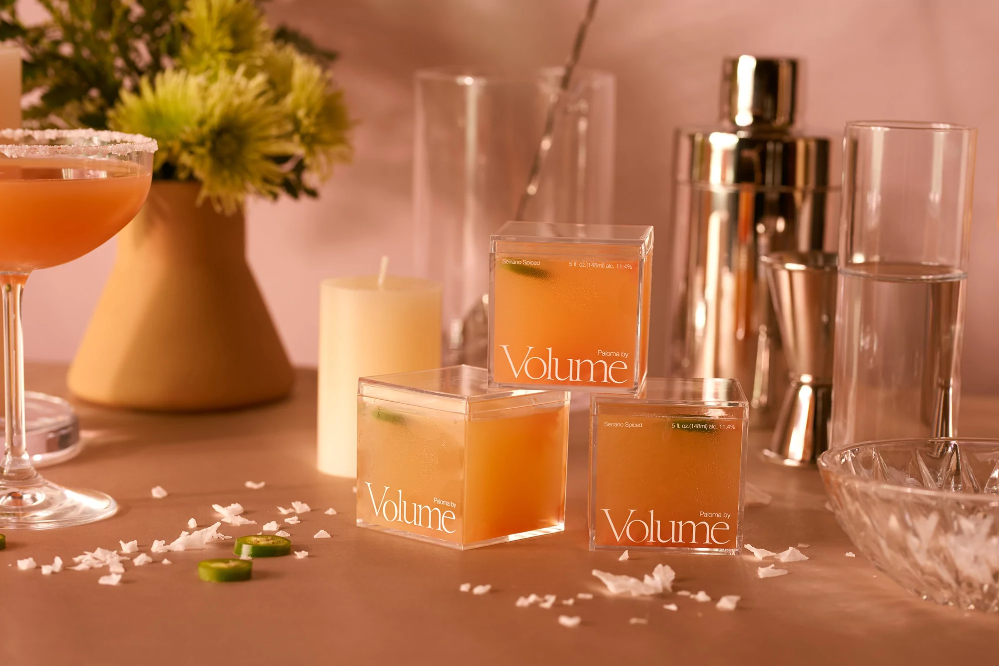 Three clear square cocktail containers filled with orange liquid, arranged on a table with a cocktail glass and bar accessories.