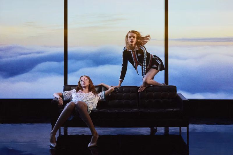 Two fashion models strike poses on a black couch in front of a window overlooking a dreamy cloudscape. The scene is lit to enhance the surreal atmosphere.