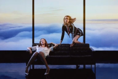 Two models in Casablanca pre-fall collection pose on a black leather sofa before a large window overlooking clouds.