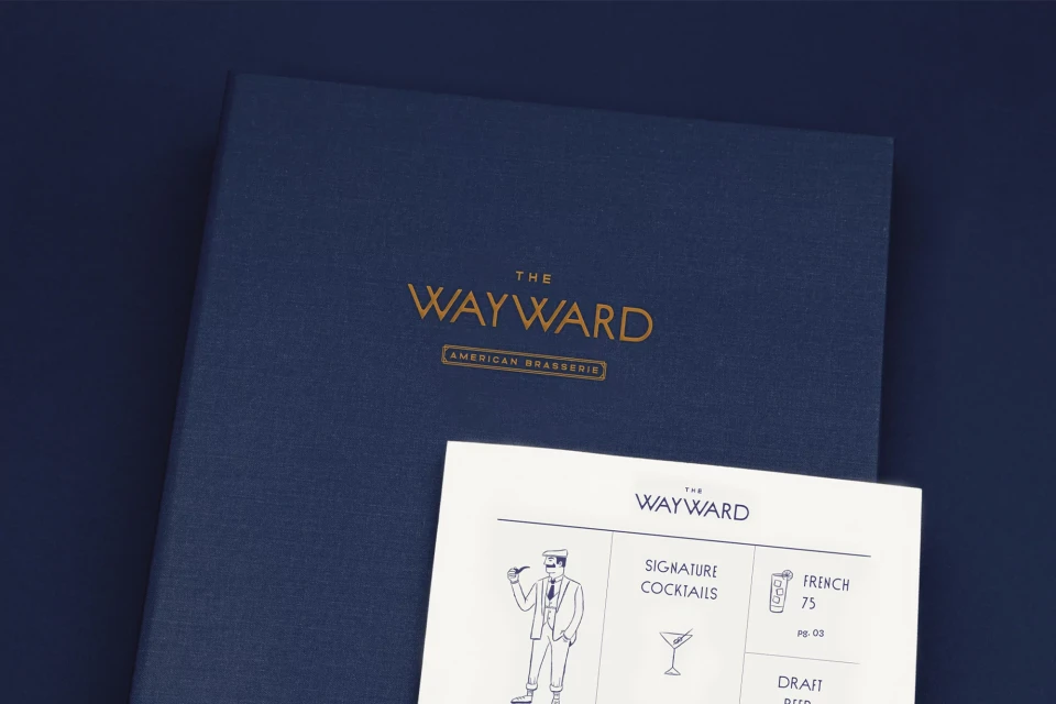 Dark blue menu book with 'The Wayward American Brasserie' in gold text, next to a white paper menu with blue line drawing.