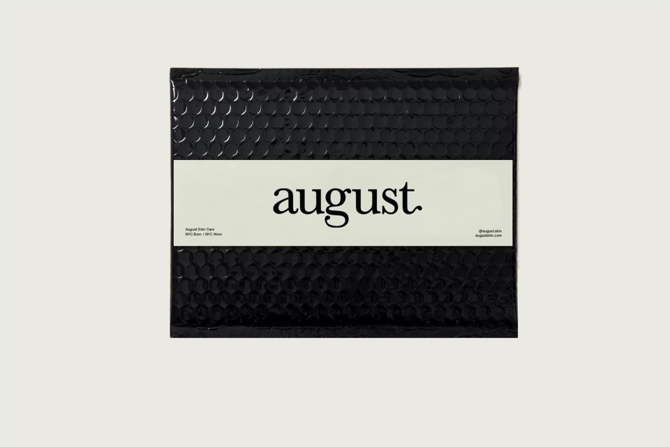 Close-up studio shot of a black August brand padded envelope with a white label, presented on a clean white background.