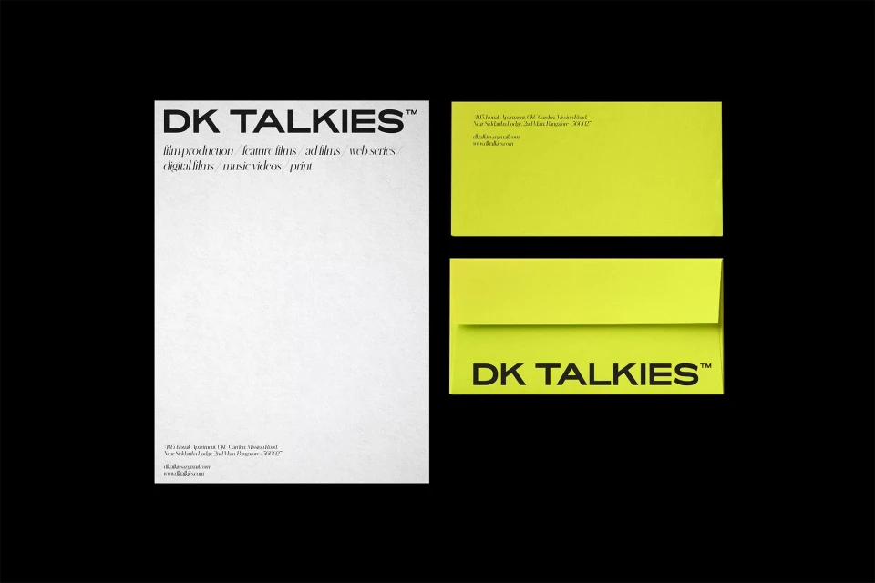 Minimalist design featuring a white poster with 'DK TALKIES' branding and film production details, alongside bright yellow envelopes on black backdrop.
