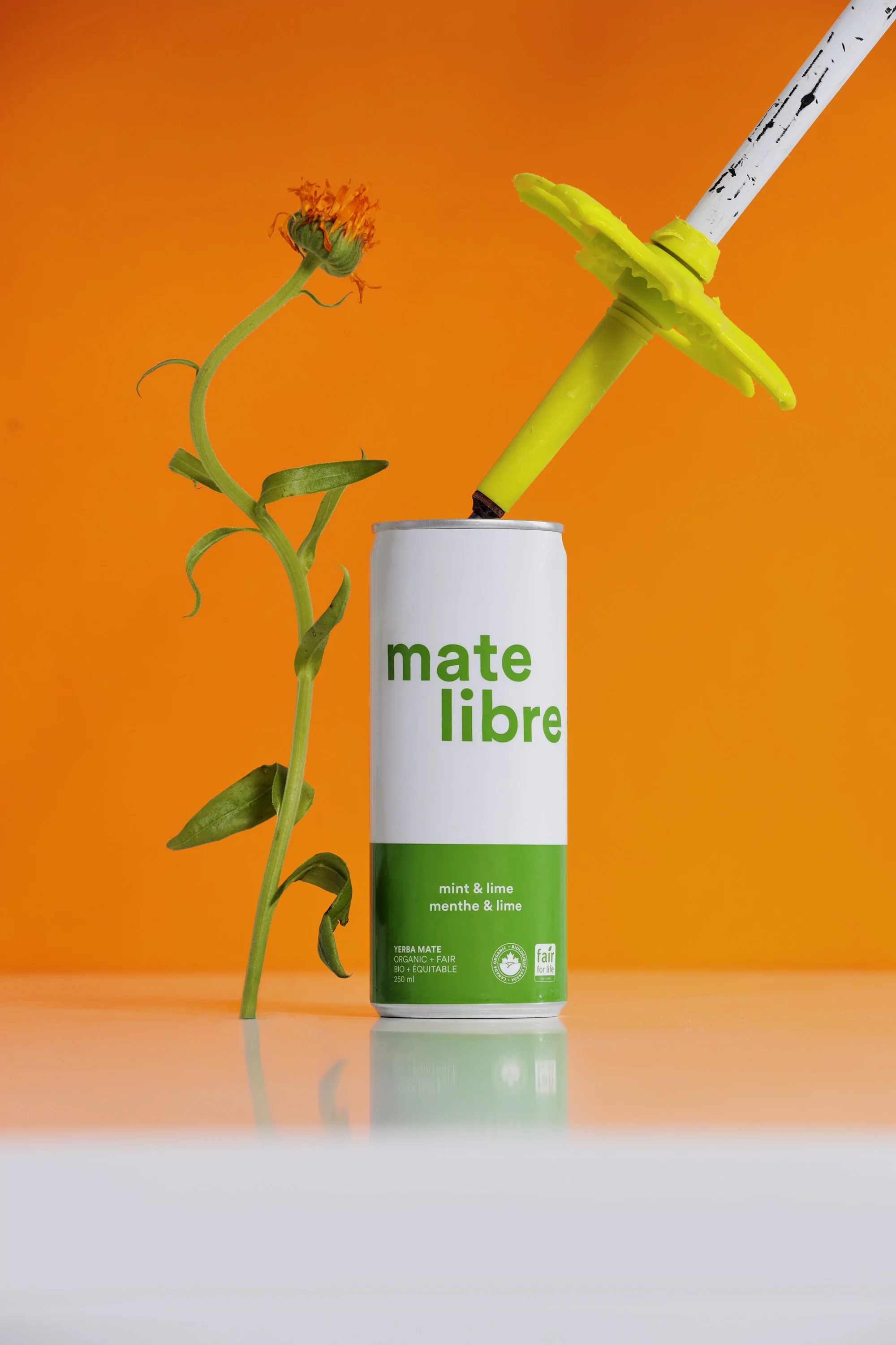 A can of Mate Libre drink with a flower and ski pole straw set against a vibrant orange backdrop in a studio setting.