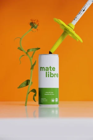 A can of Mate Libre drink with a flower and ski pole straw set against a vibrant orange backdrop in a studio setting.