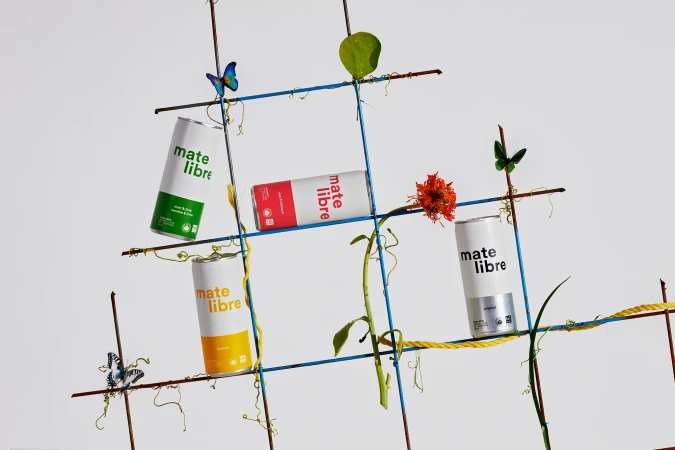 Four cans of Mate Libre drink are arranged on a blue grid with flowers and vines against a white background in a studio shot.