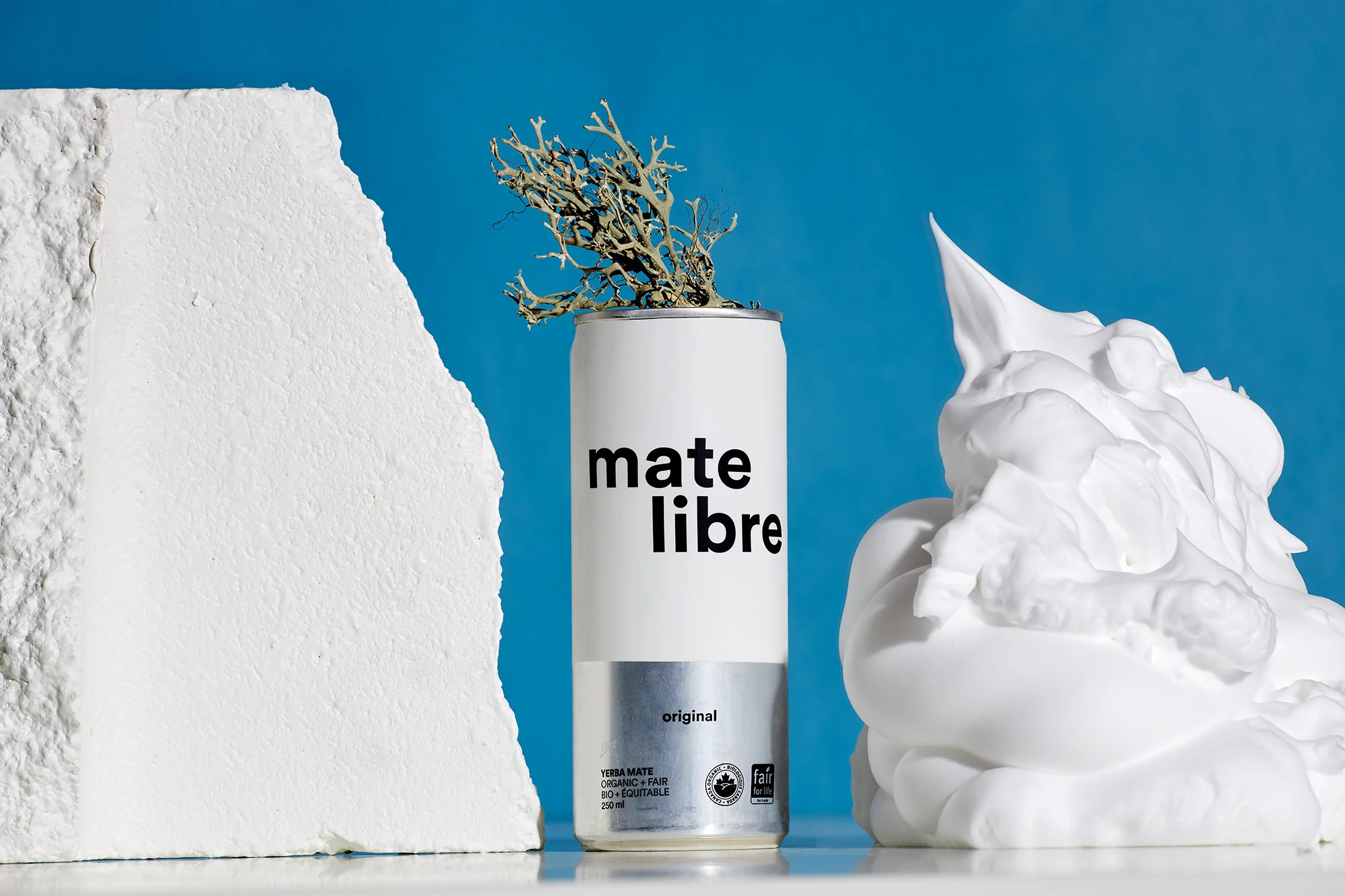 A white can of Mate Libre yerba mate sits among decorative objects against a blue background in a minimalist still life.