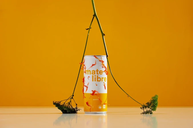 A can of Mate Libre beverage with Passion fruit flavor, staged with thin leafy stems against a solid yellow background. Studio product shot.