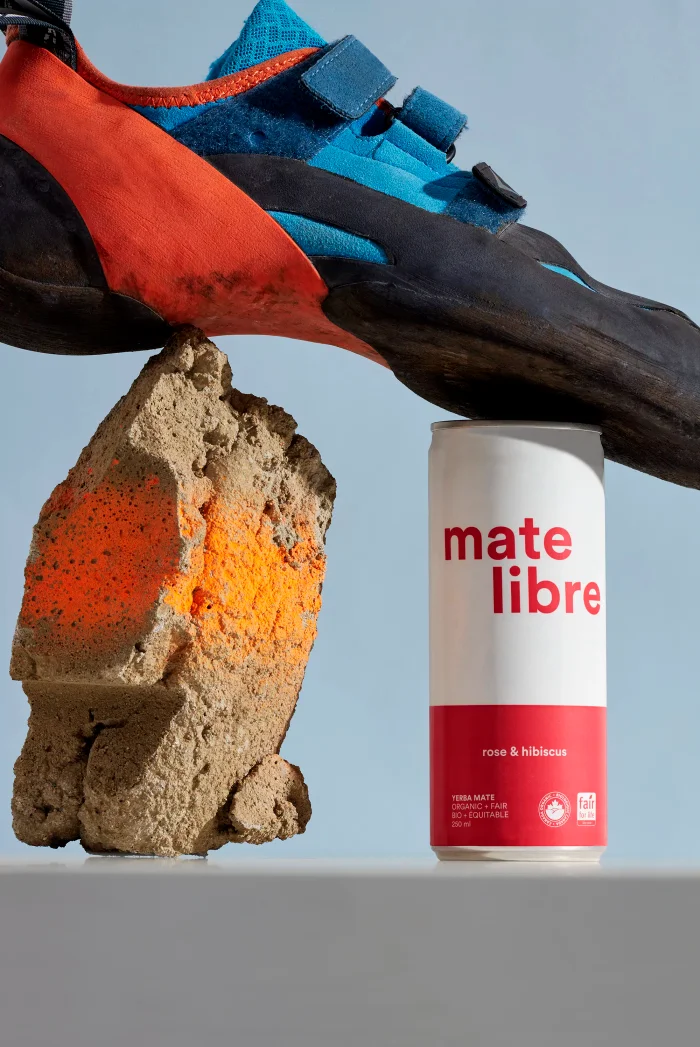 A climbing shoe resting on a textured rock and a Mate Libre beverage can against a solid blue background