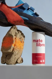 A climbing shoe resting on a textured rock and a Mate Libre beverage can against a solid blue background