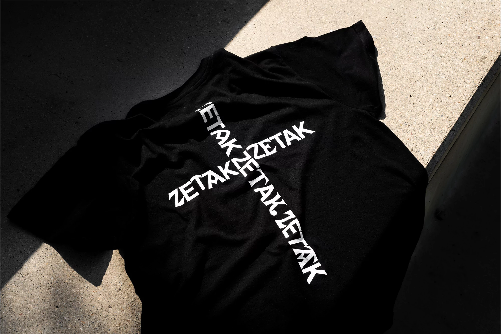 Black t-shirts are stacked on a concrete surface with some bright lighting accentuating the graphic white cross design on the shirt.