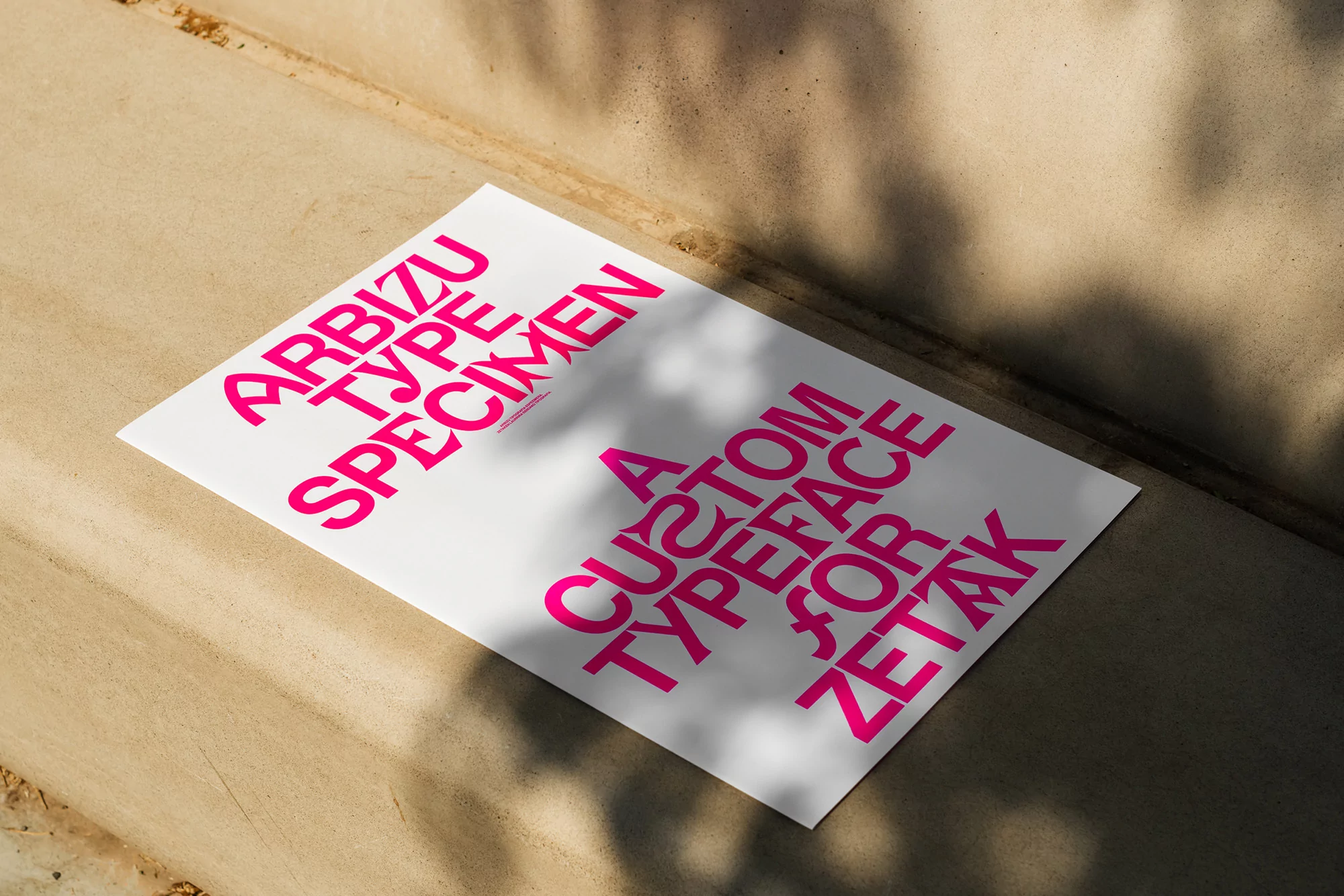 Arbizu type specimen poster in bright pink lettering, lying on a concrete surface, with leaf shadows creating an abstract background.