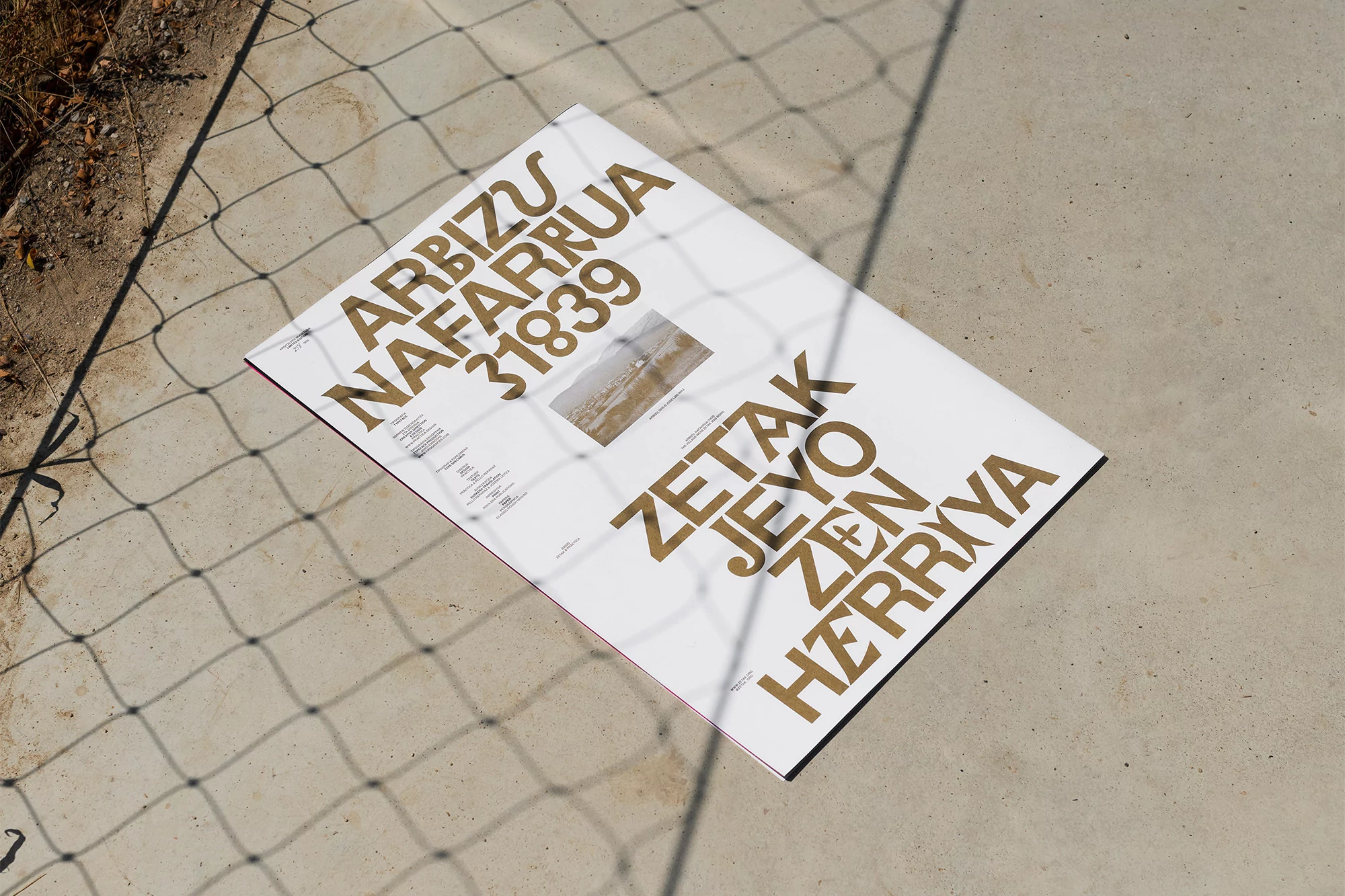 Promotional poster featuring typography in gold tones on a white background, resting on a grey surface with shadow of a net.