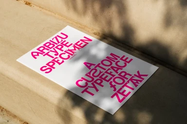 White poster featuring bold magenta typographic specimen text resting on a concrete surface with dappled light